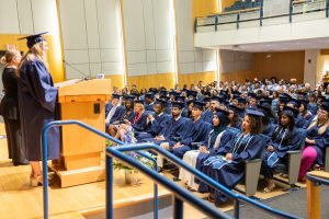 A woman in graduation regalia stands on stage addressing an audience of students also in regalia.
