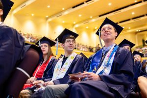 Three students in graduation regalia sit and listen to a speaker.