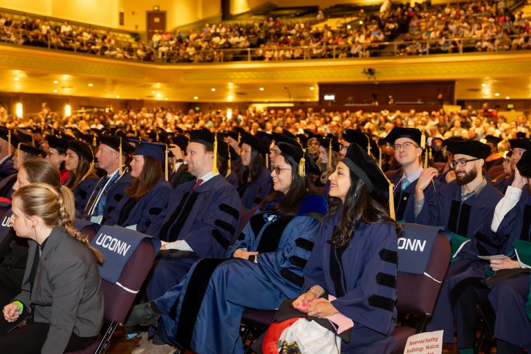 A row of people in commencement regalia sit in an auditorium.