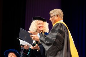 A woman and man in commencement regalia stand side by side as the man examines an honorary diploma.