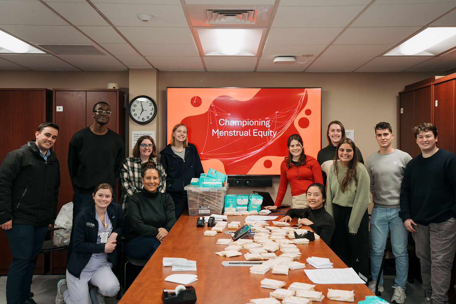 group portrait of 12 in conference room with menstrual products on table in front of "Championing Menstrual Equity" sign