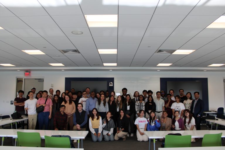 A large group of students and faculty pose together in a modern classroom with white walls and ceiling tiles.