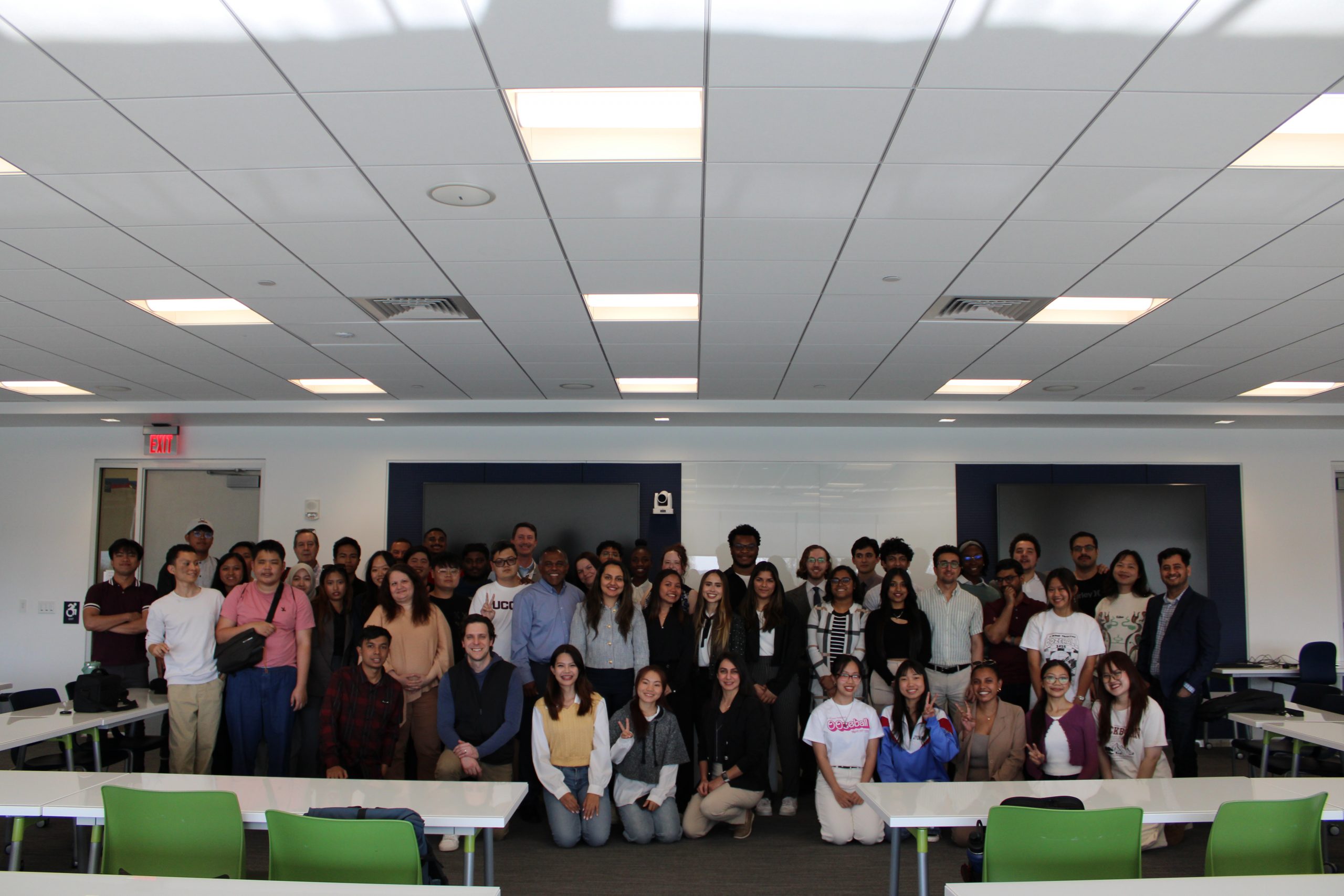 A large group of students and faculty pose together in a modern classroom with white walls and ceiling tiles.