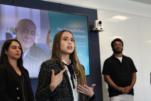 A woman speaks during a classroom presentation with a photo of her grandfather displayed on the screen behind her, flanked by two other presenters.