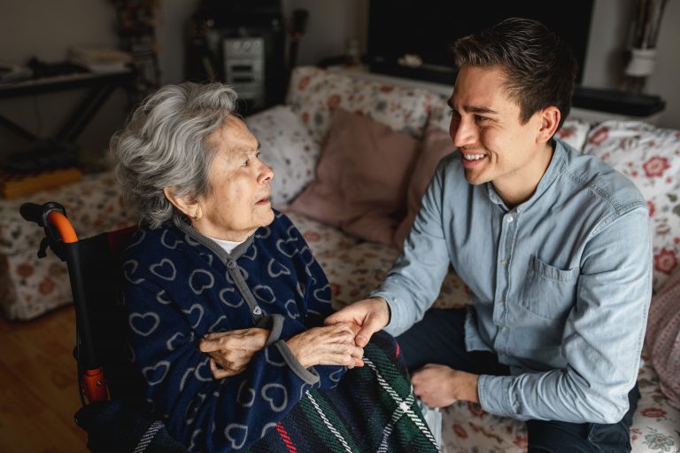 Young man sitting next to an old sick aged woman in wheelchair taking her hands while talking and smiling.