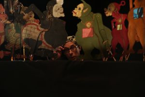 A man with glasses peers through a stage of puppets during a performance.