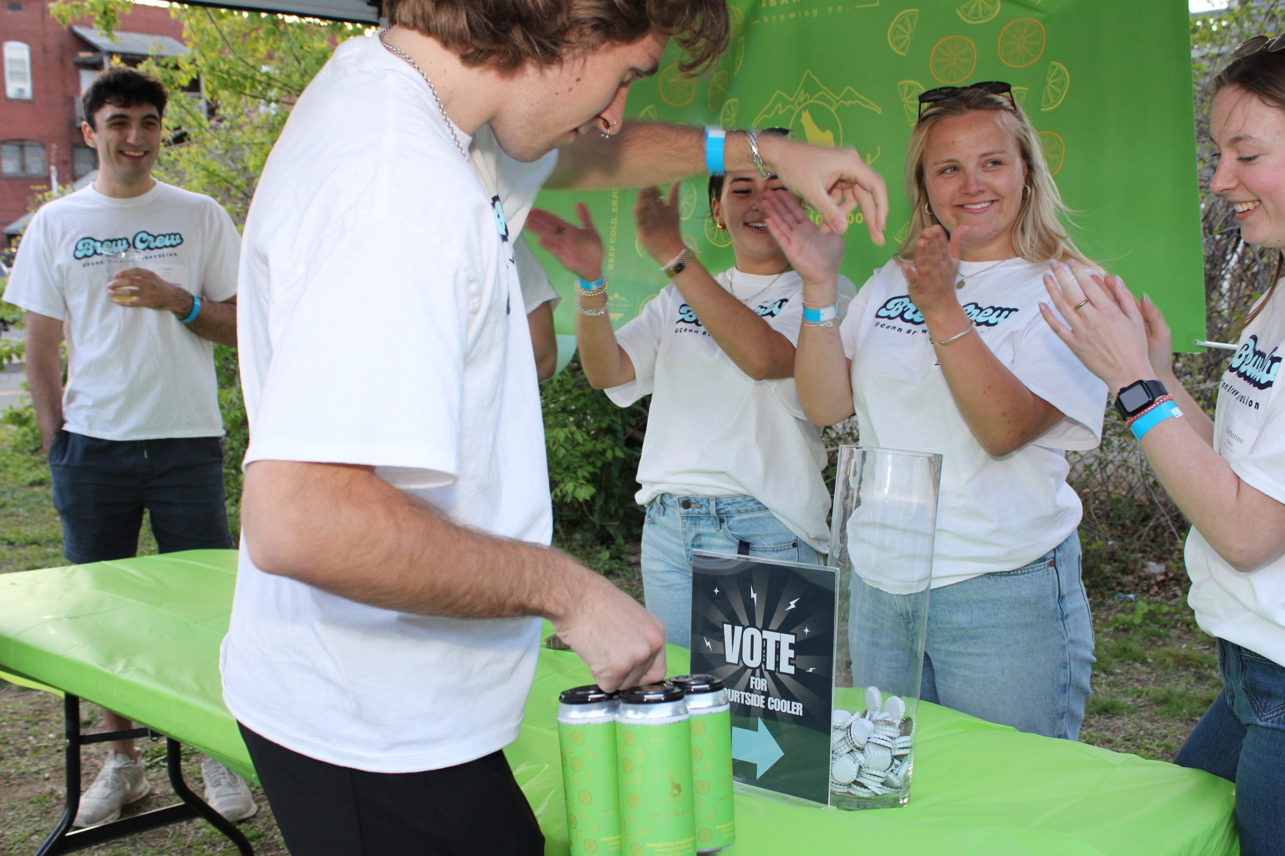 A man in a while shirt drops a bottle cap into a plastic receptacle while three women in white shirts applaud.