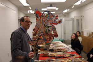 A man holds a stick attached to a multicolored two-dimensional puppet while students sitting around a table look on. Other puppets are stacked on the table.