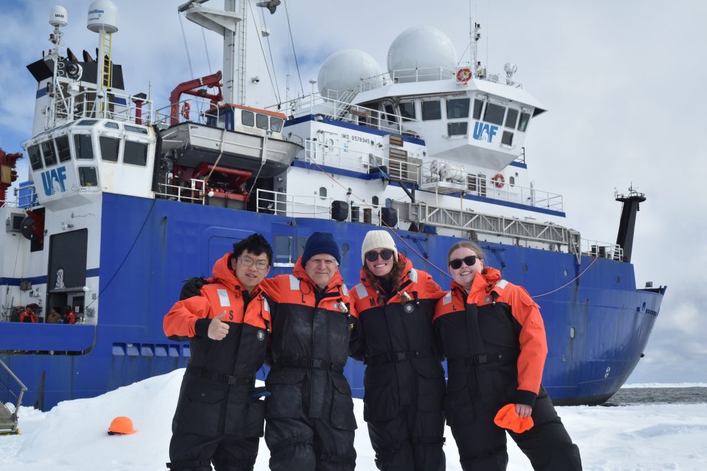 Four researchers stand outside of a large blue and white research boat.