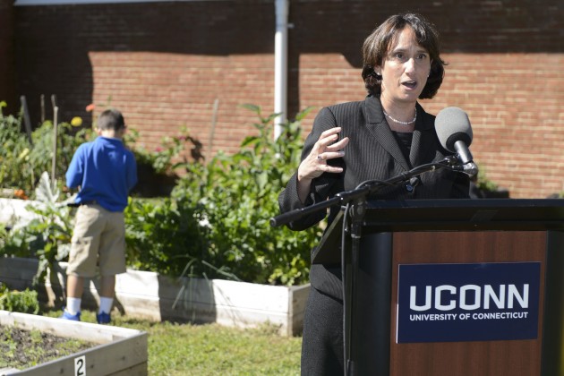 A woman in a suit speaking at a podium with the UConn logo while children explore a garden in the background