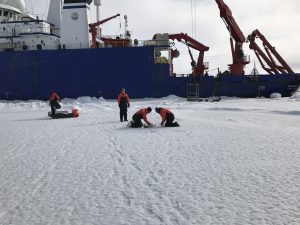 Four people dressed in black and red snow gear take samples from sea ice close to a large blue research boat.