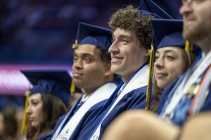 Students in graduation regalia listen to a speaker.