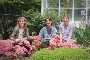 Three women sitting in a flower bed surrounded by green bushes and pink flowers