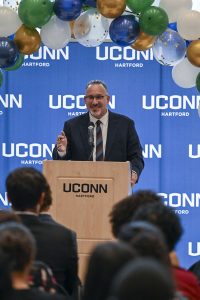 Former US Education Secretary Miguel Cardona speaks at a podium with a blue backdrop that says "UConn" behind him.