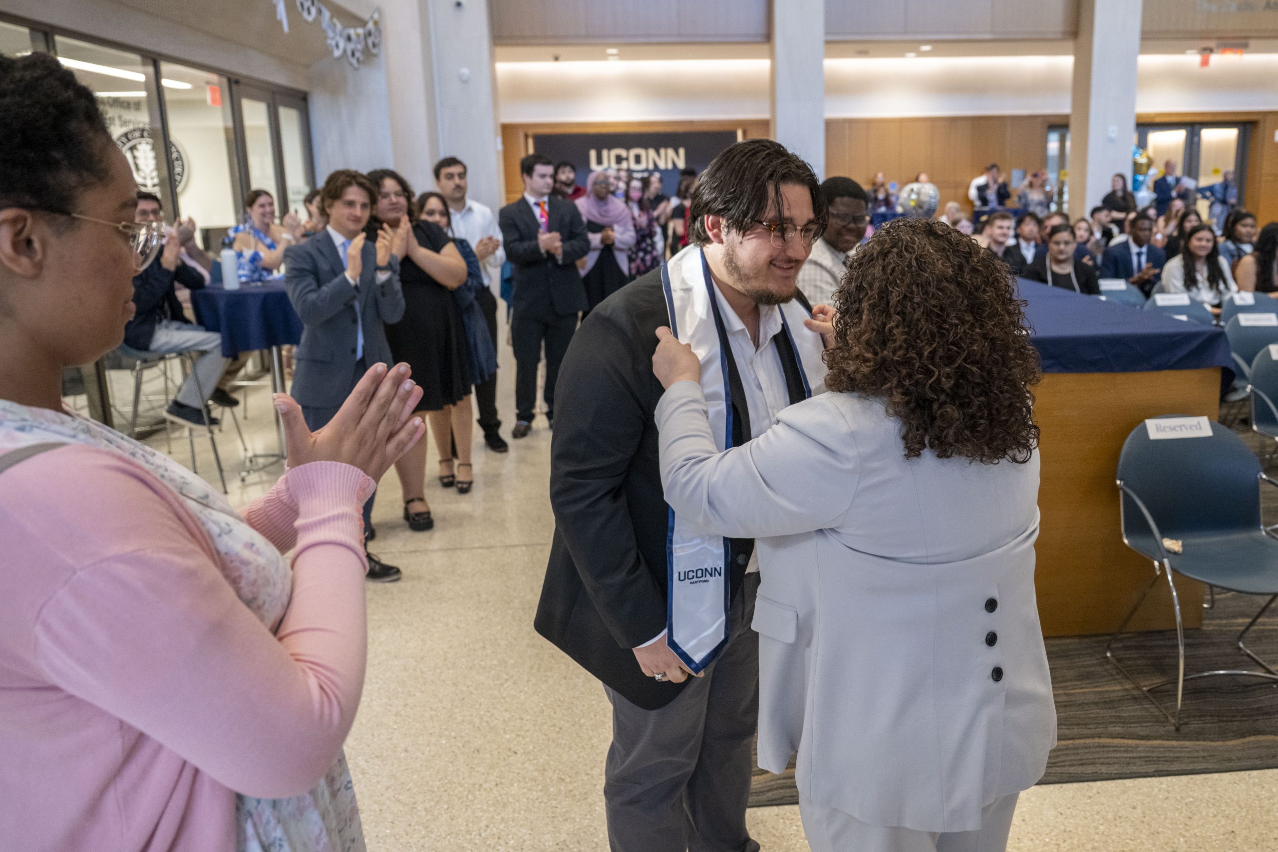 A woman in grey with her back to the camera puts a white and blue stole around the neck of a student in a dark blazer.