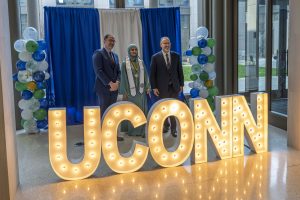 Two men in suits stand beside a young woman in green, posing together behind waist-high illuminated letters that spell UConn.