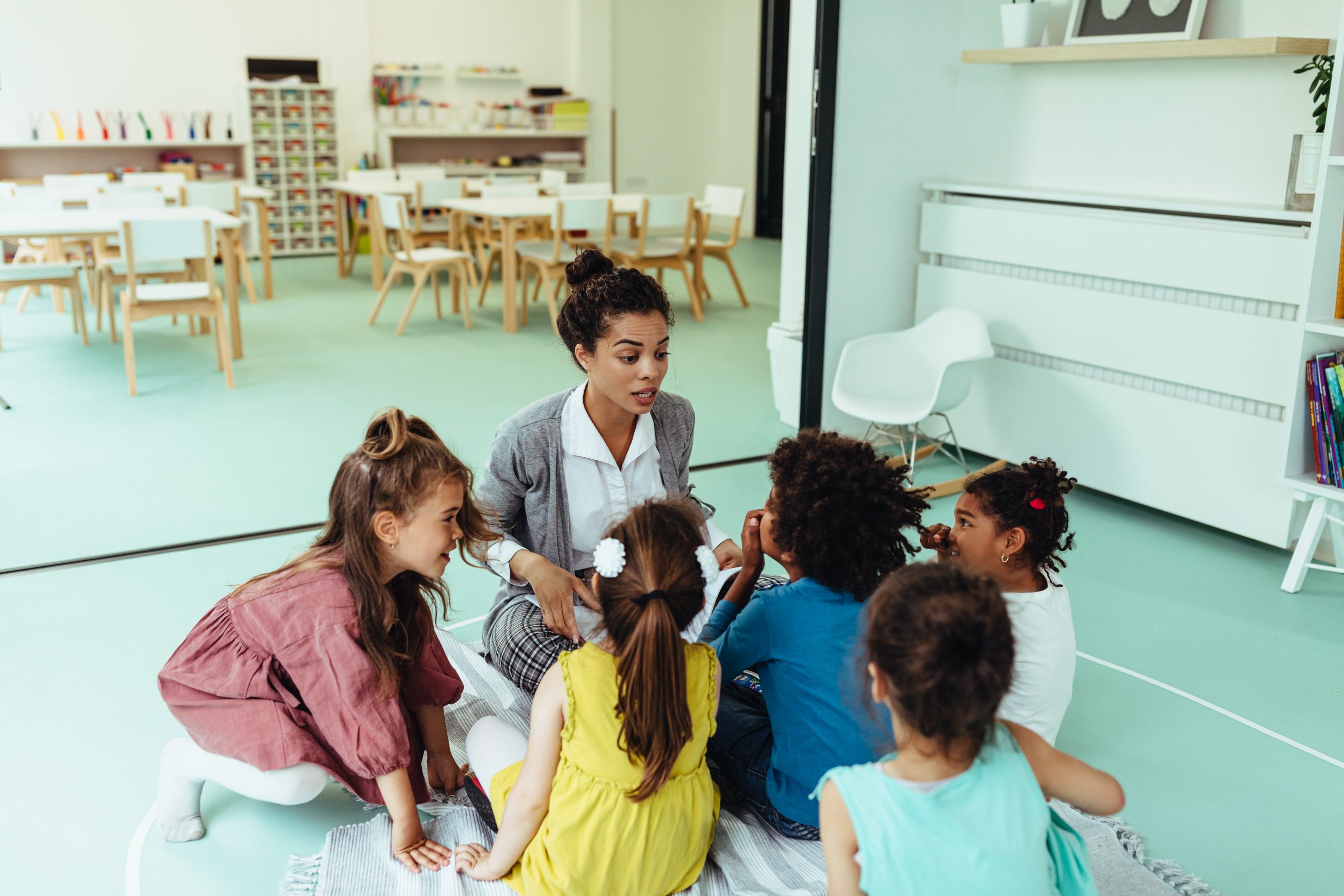 A teacher kneels on the floor of a classroom, surrounded by young students.