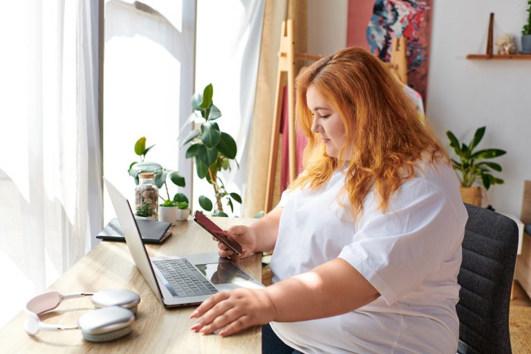 A woman with excess body weight sits at a desk and uses a smartphone and laptop