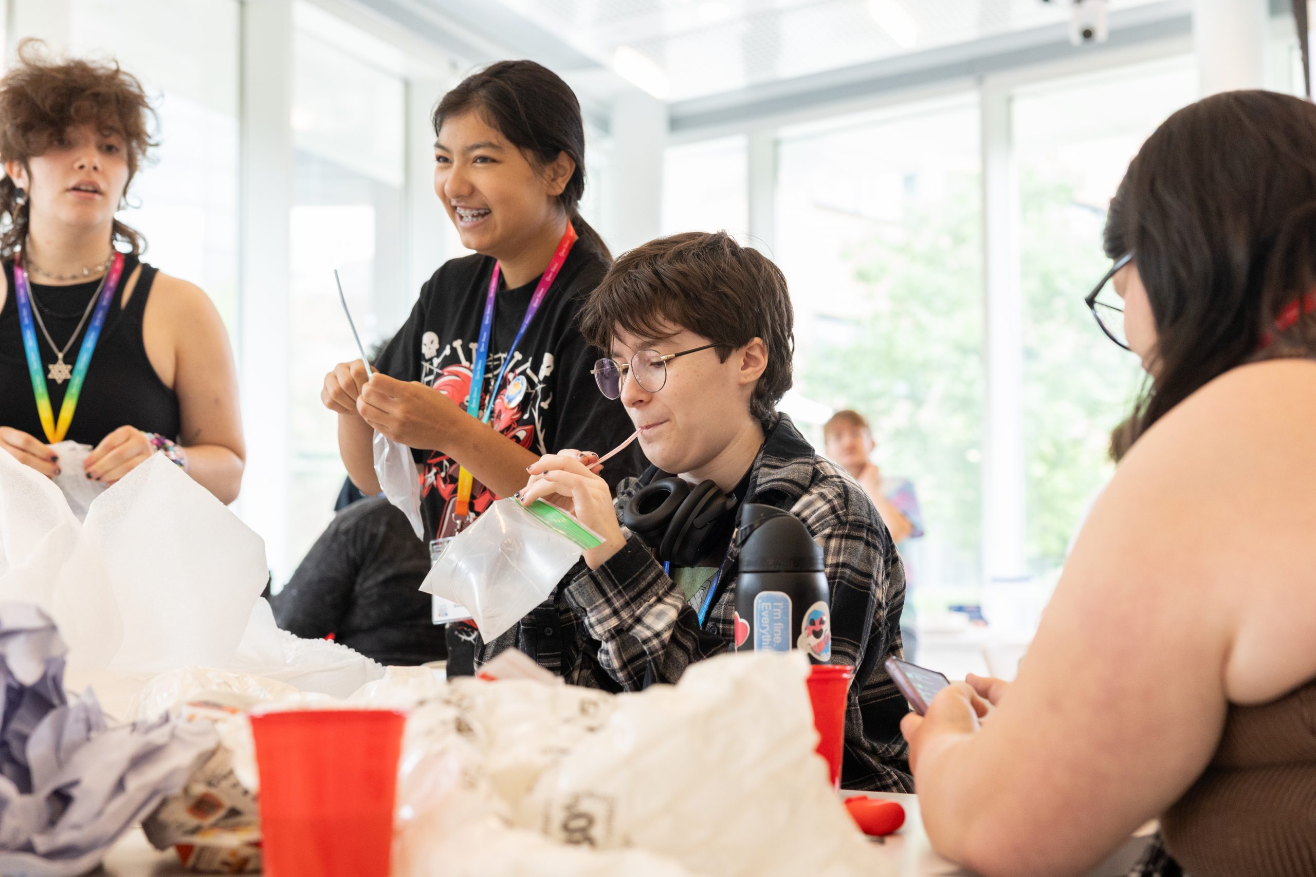 Four students wearing rainbow lanyards use materials like plastic cups and baggies to build contraptions