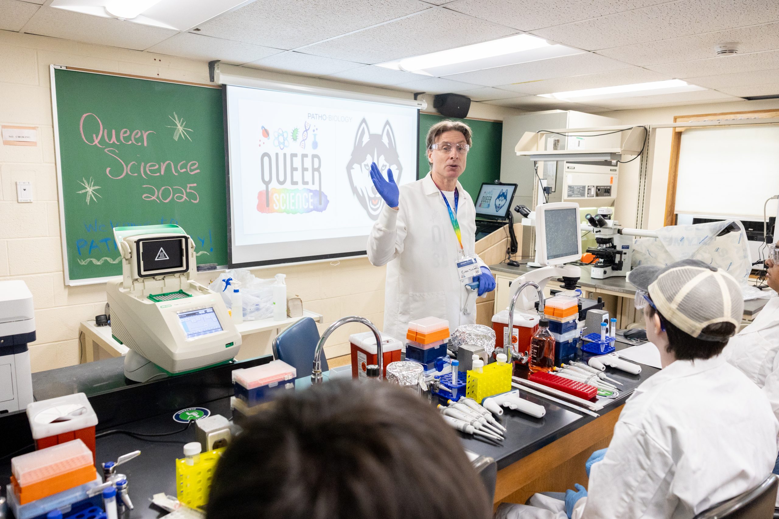 A professor wearing a labcoat and rainbow lanyard lectures to a small lab classroom full of students, with "Queer Science 2025" written on the chalkboard in the background
