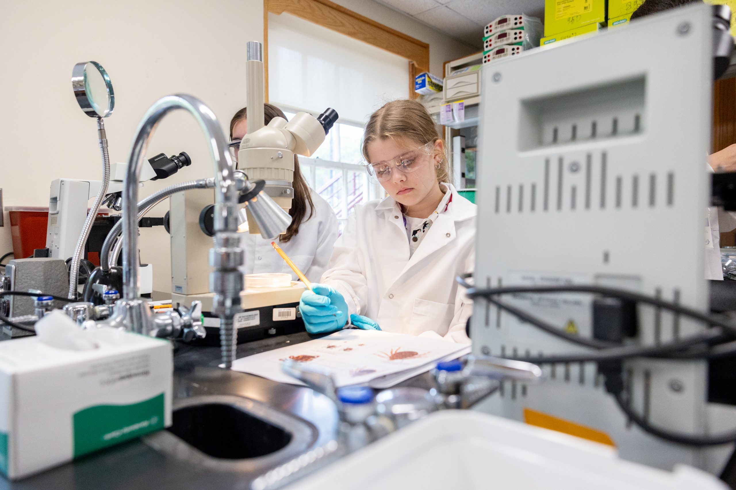 A young person wearing a labcoat and goggles examines a worksheet with photos of different kinds of ticks