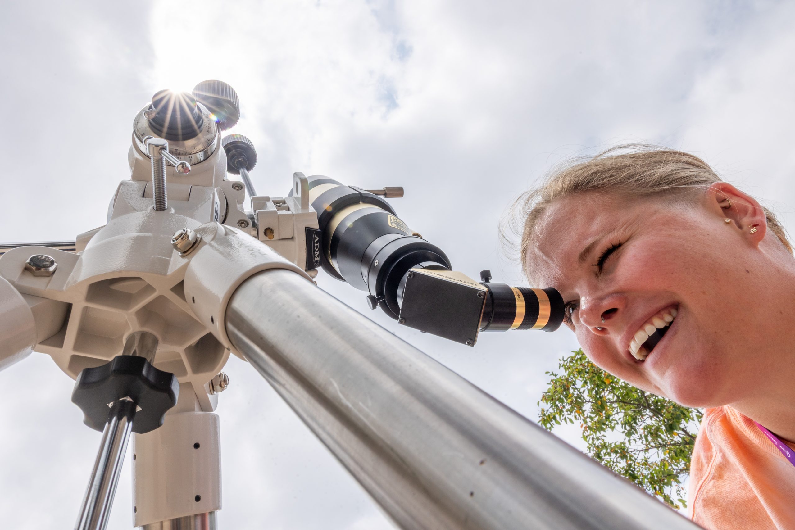 Sun glints off a telescope pointed toward the daytime sky as a person smiles and looks into the eyepiece