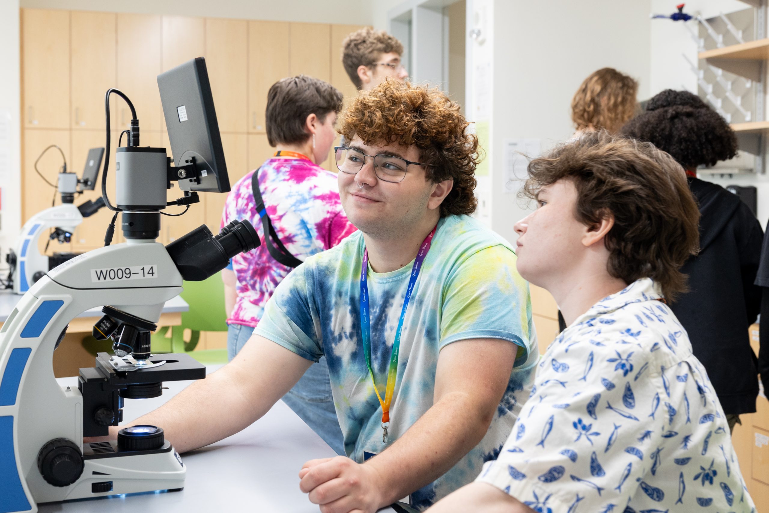 A person in a tie-dyed t-shirt helps a younger student examine the output images from a microscope