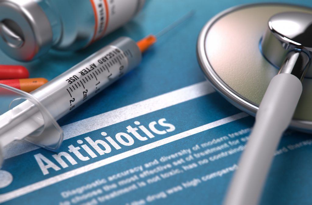 Medical implements, including a syringe and stethoscope, lie across a paper with the word "antibiotics" written on it.