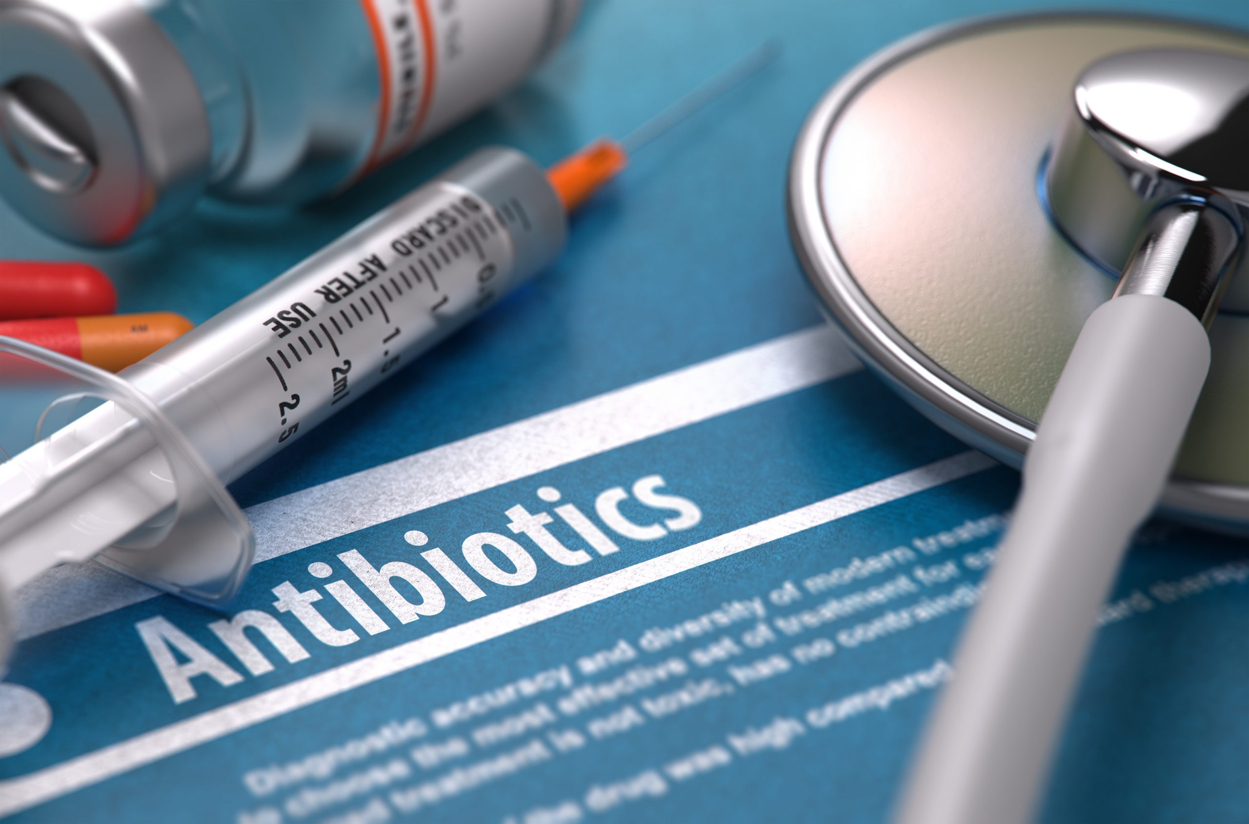 Medical implements, including a syringe and stethoscope, lie across a paper with the word "antibiotics" written on it.