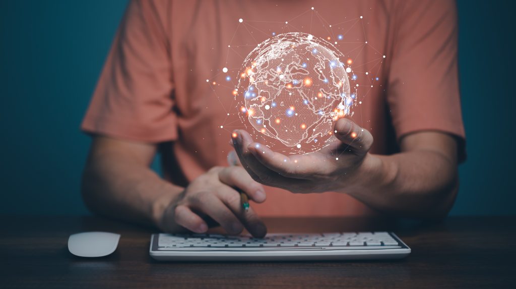 A man sitting in front of a keyboard holds a holographic image of the globe in his hand.
