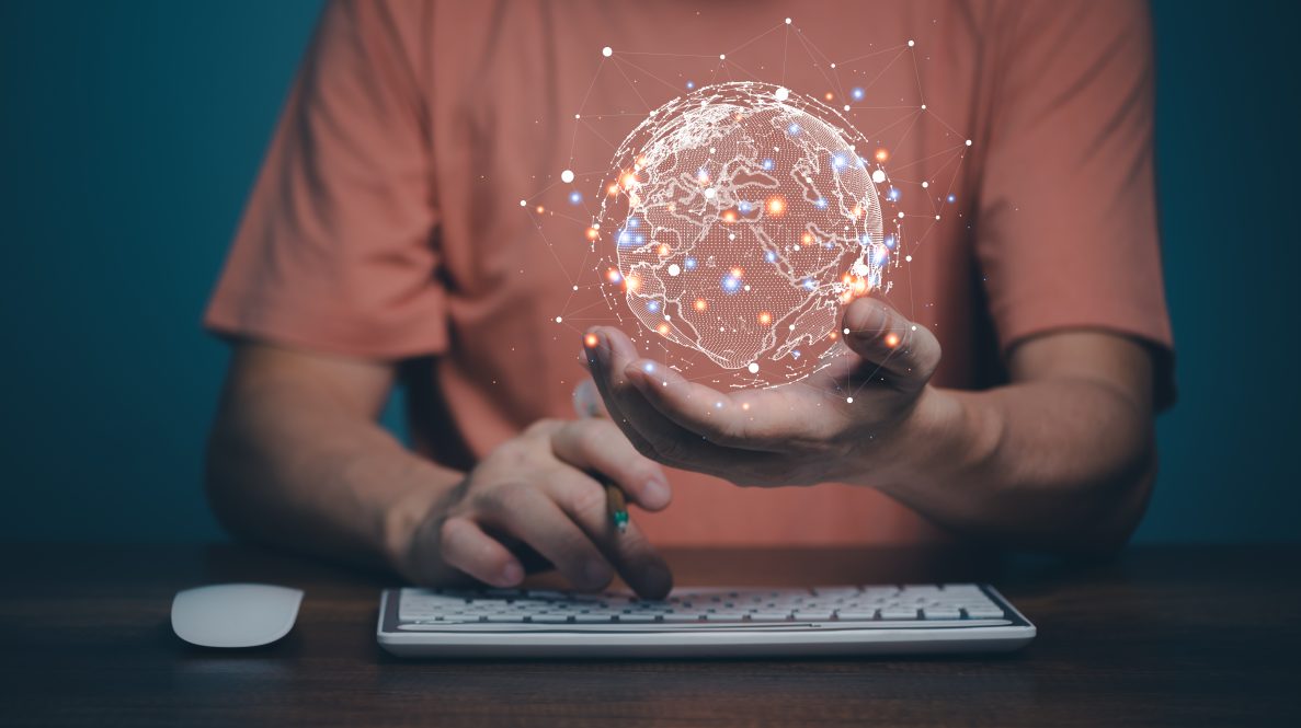 A man sitting in front of a keyboard holds a holographic image of the globe in his hand.