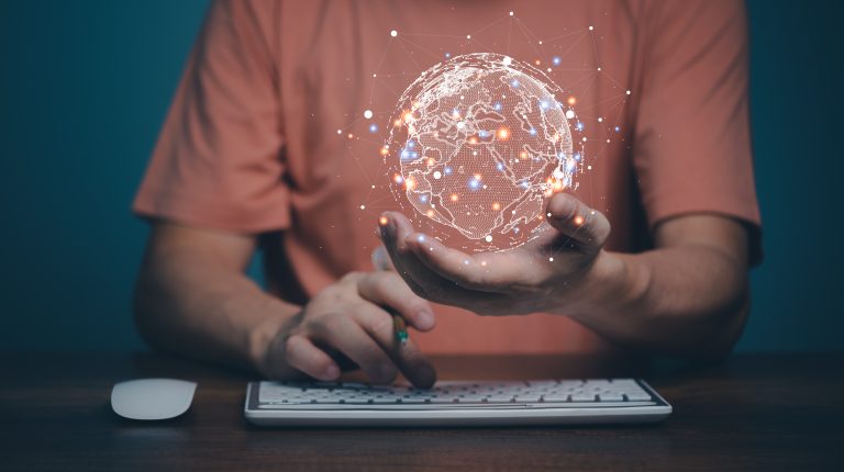 A man sitting in front of a keyboard holds a holographic image of the globe in his hand.