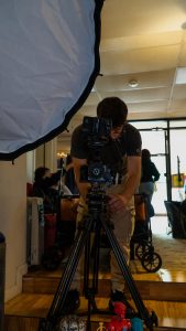Evan Elmore, a dark haired young man wearing glasses and a gray t-shirt, handles a digital video camera, while three other young people hang out in the background.