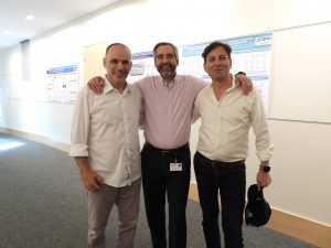Three men in button-down shirts and slacks smile for a photo. The man in the middle has his arms around the two on the ends