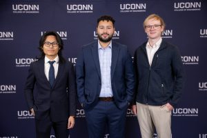 Three men posing in front of a UConn College of Engineering backdrop, two wearing suits and one in casual business attire.