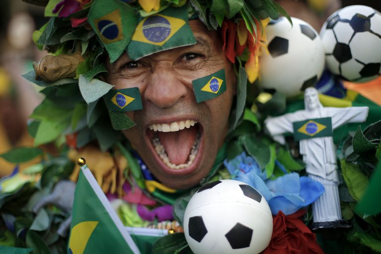 A fan of Brazil celebrates with soccer balls around his face, and his face painted with the Brazil flag