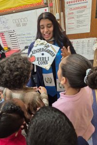 A female college student holds up a decorated mortarboard to show four elementary school students.