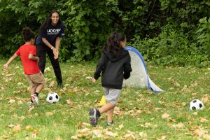 A female college student plays soccer outside with two elementary school students.