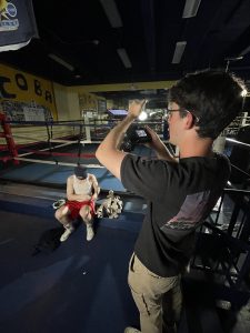 A young man shoots video with a hand-held digital camera while his subject, another man wearing a white tank top and red shorts, sits outside a boxing ring.