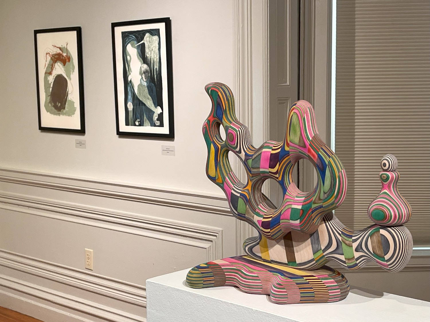 A multicolored, wooden sculpture sits on a pedestal in an art gallery with other framed art in the background.