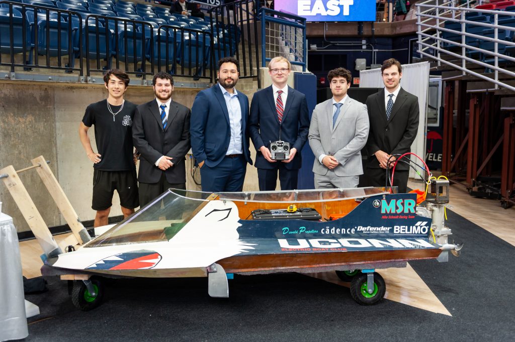Six students posing with a small race boat at an indoor event, some in suits and one in casual clothes.