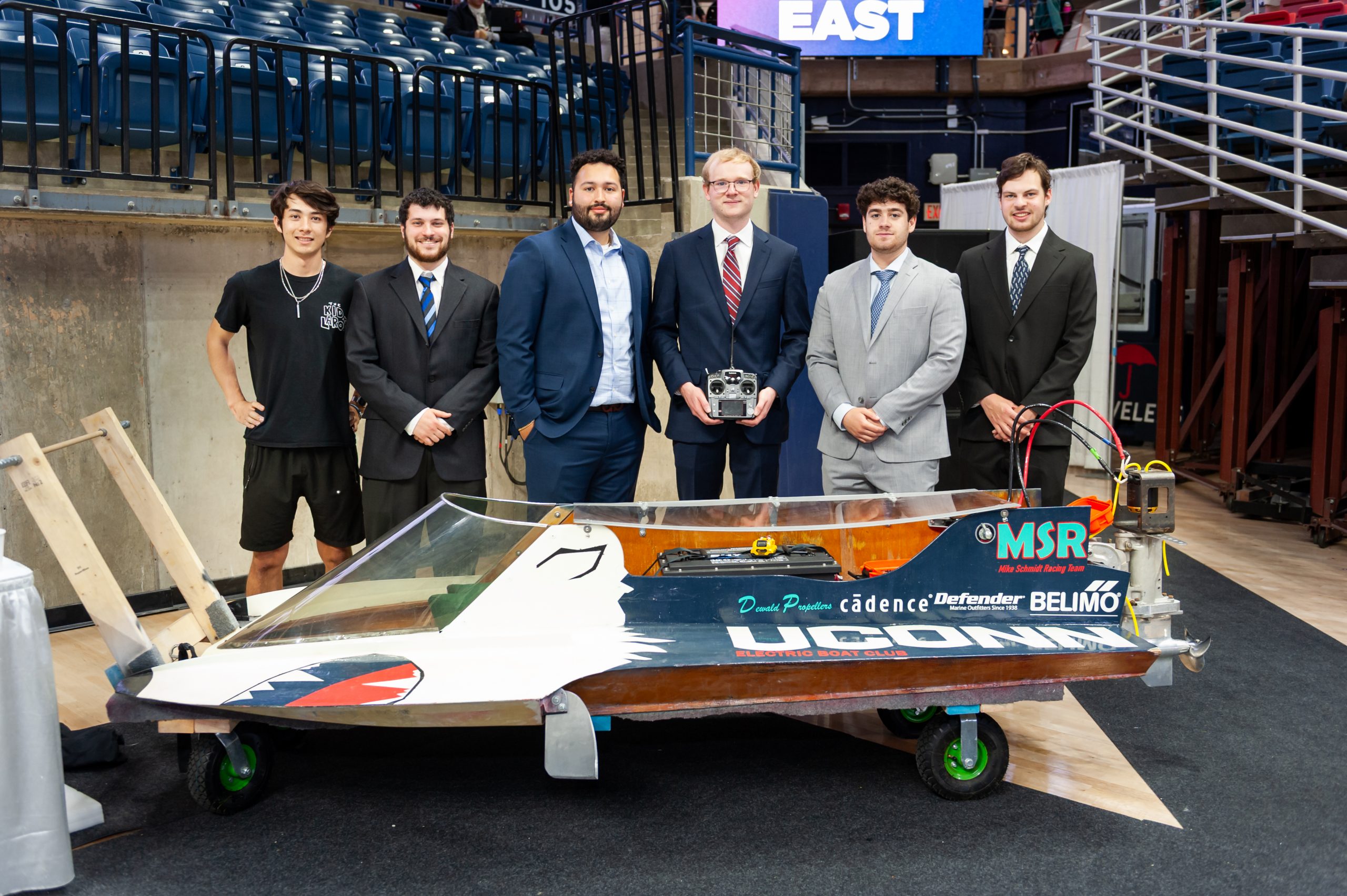 Six students posing with a small race boat at an indoor event, some in suits and one in casual clothes.