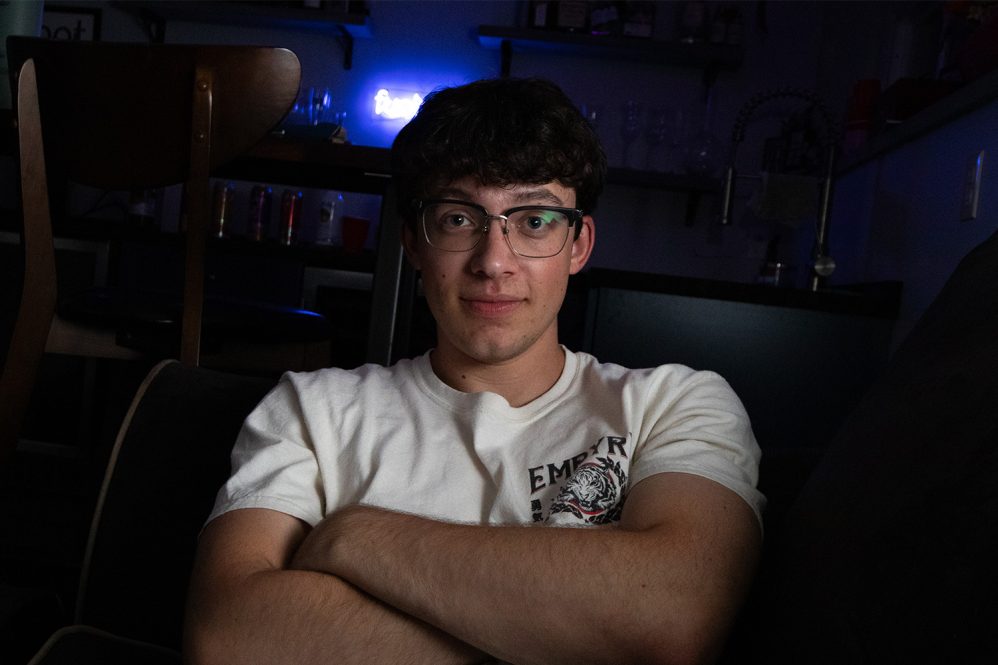 Evan Elmore, a young man with dark hair and glasses, wears a white t-shirt and sits with his arms crossed in this portrait.