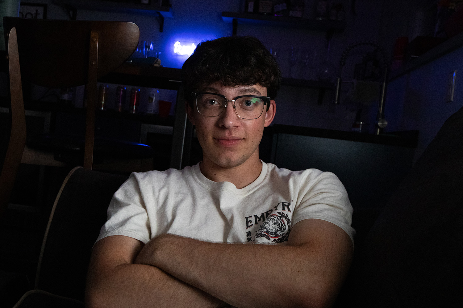 Evan Elmore, a young man with dark hair and glasses, wears a white t-shirt and sits with his arms crossed in this portrait.