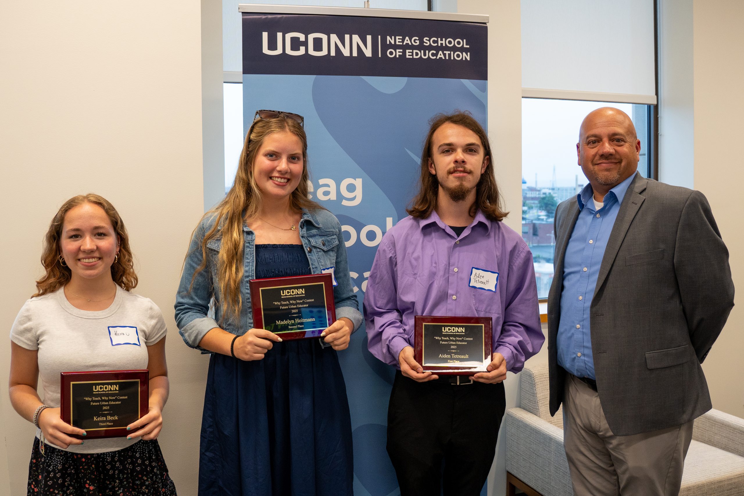 Three high school students - two female, one male - stand next to Dean Jason G. Irizarry in front of a light blue vertical banner that reads "UConn Neag School of Education." Each of the three students holds a small wood and metal award plaque.