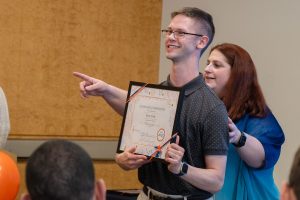 man holding certificate, woman behind him directing him toward a camera