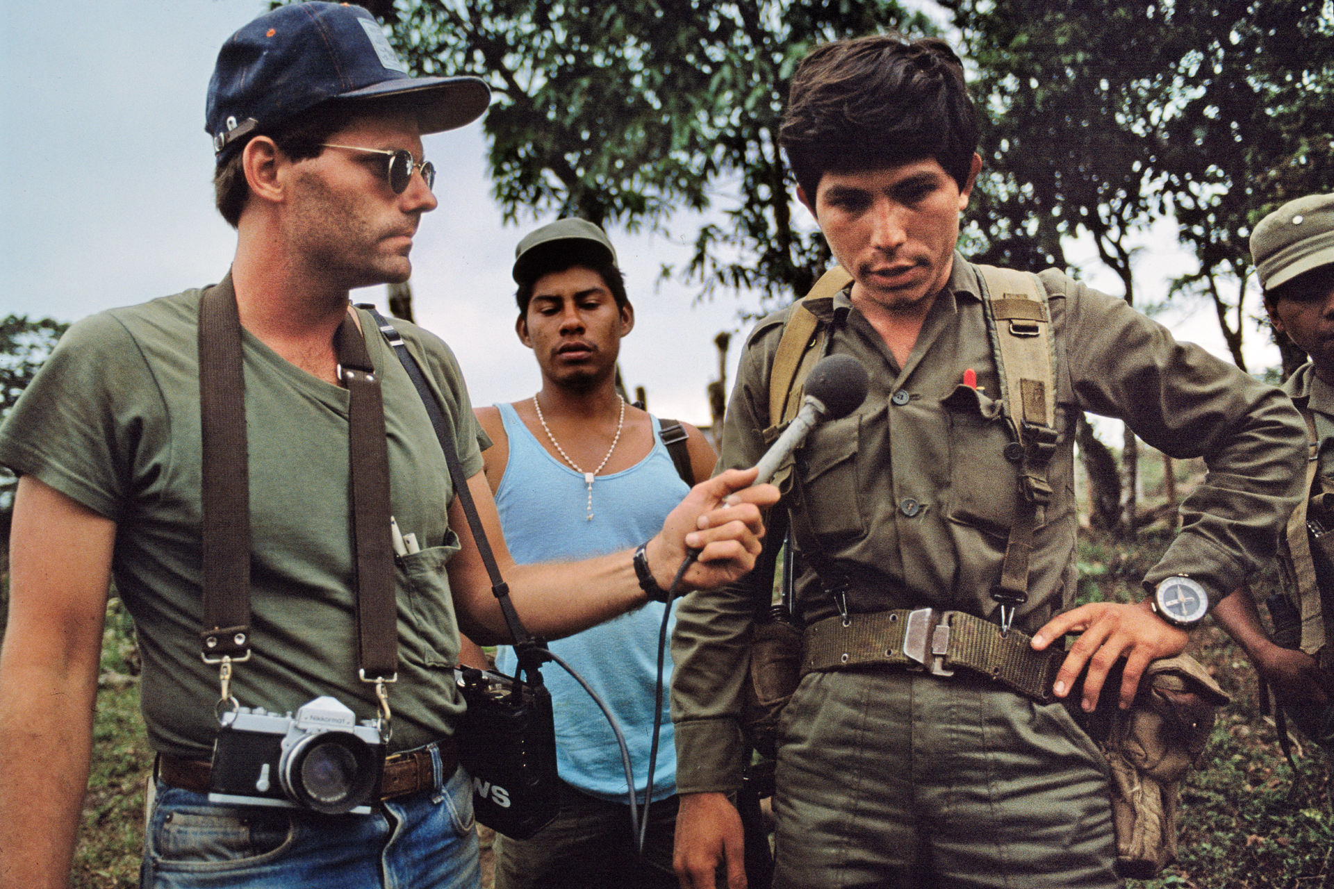 A reporter with a camera hanging from his neck and microphone in his hand interviews a military officer.