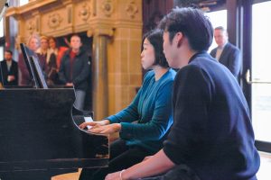 Two people sit at a piano playing while onlookers watch