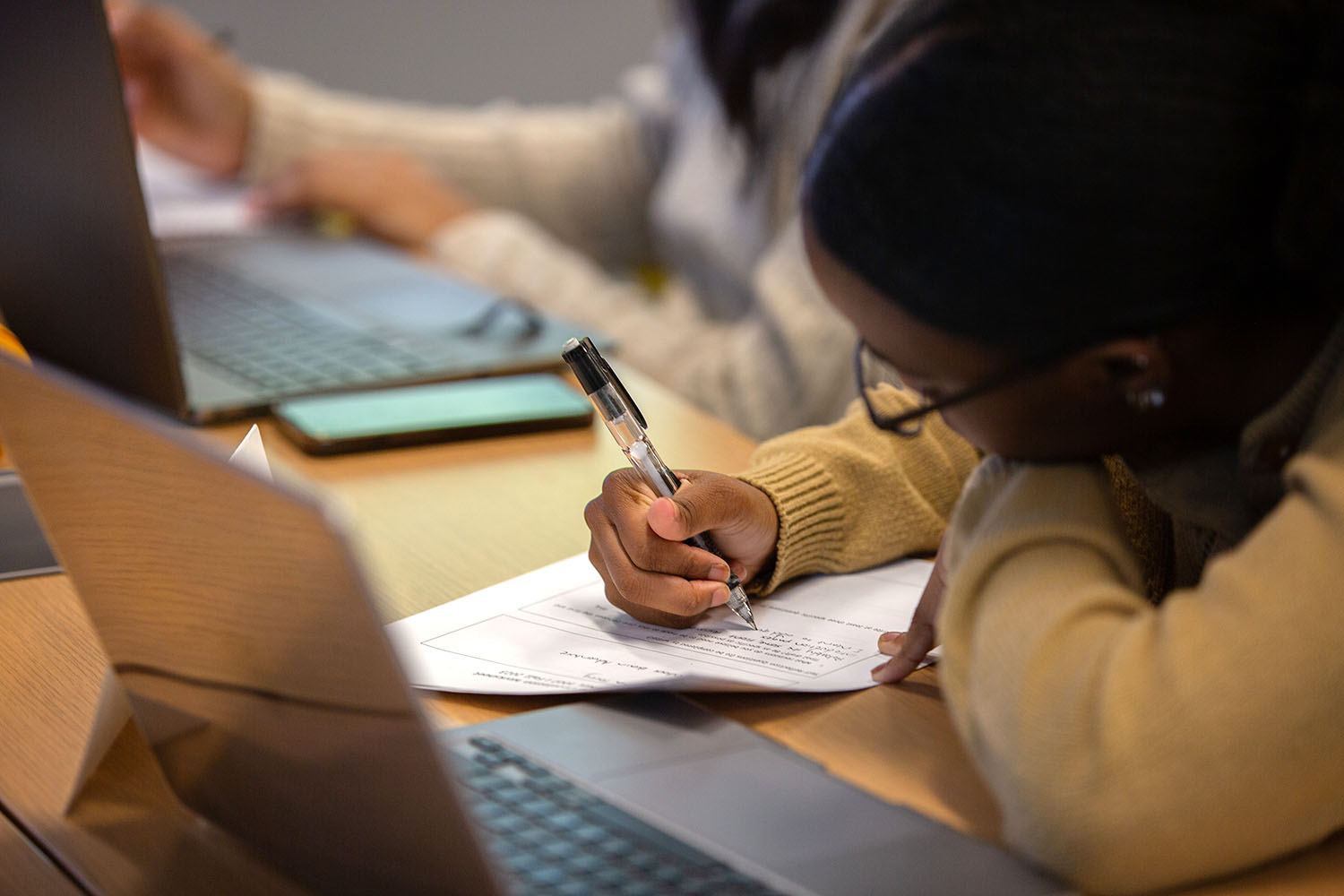 A student focuses on completing a worksheet with a pen during class, with a laptop open in front of them.