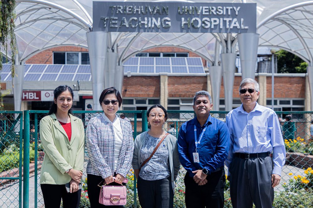 Group portrait of five outdoors in front of "Tribhuvan University Teaching Hospital" sign